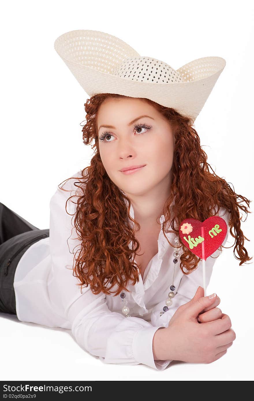 Young pretty caucasian girl wearing big hat and holding heartlike bon-bon candy dreaming about summer time and vacation isolated on white background. Young pretty caucasian girl wearing big hat and holding heartlike bon-bon candy dreaming about summer time and vacation isolated on white background