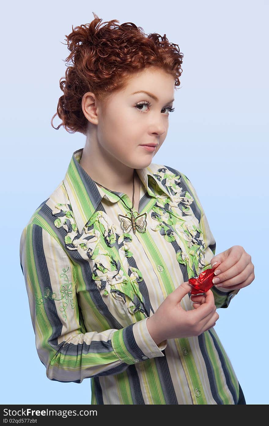 Calm and beautiful young red haired girl with curly natural hair unwraping chokolate candy bar standing with curious facial expression and isolated over light blue background. Calm and beautiful young red haired girl with curly natural hair unwraping chokolate candy bar standing with curious facial expression and isolated over light blue background