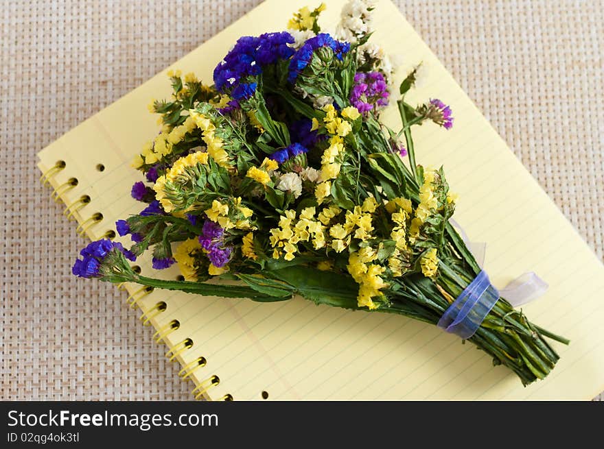 A bunch of immortelle flowers tied with violet organza ribbon set on a yellow copybook on the table. A bunch of immortelle flowers tied with violet organza ribbon set on a yellow copybook on the table