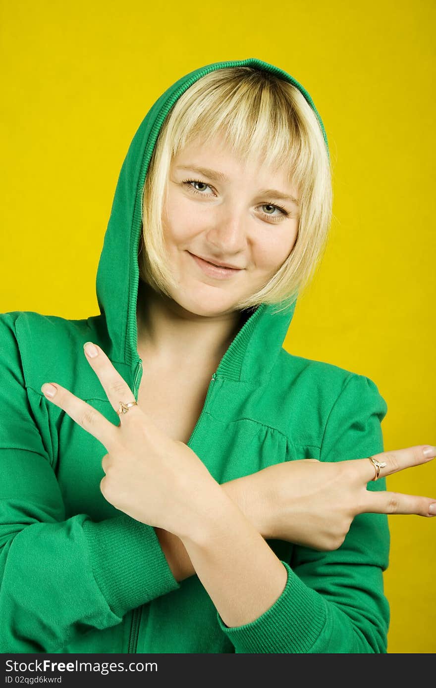 Blonde woman on a yellow background shows the sign of Peace. In the green jacket and hood. Blonde woman on a yellow background shows the sign of Peace. In the green jacket and hood