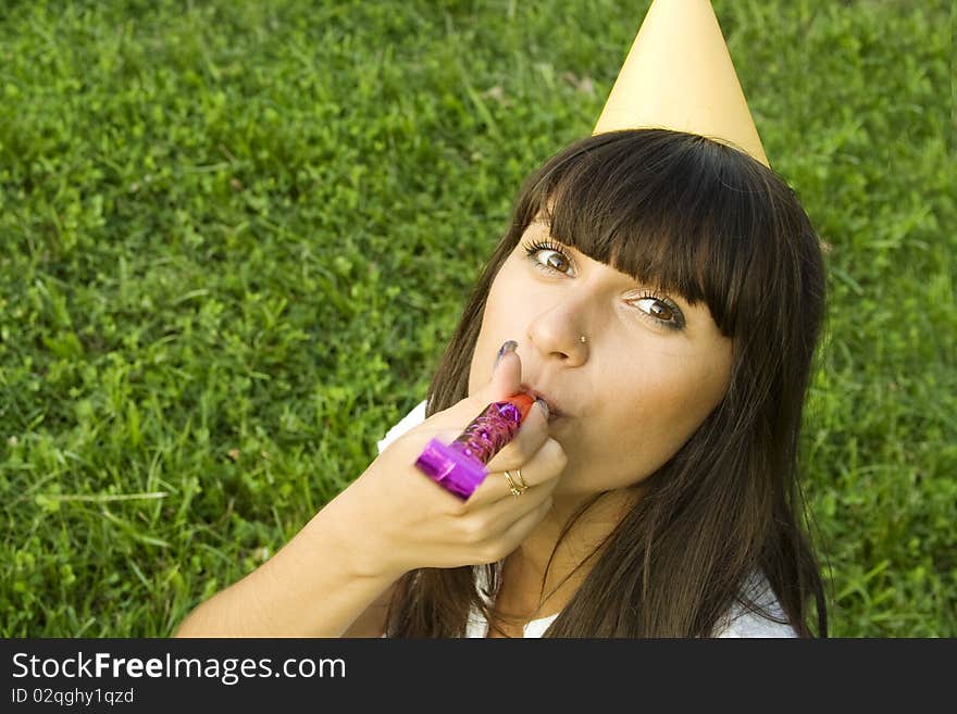 Beautiful girl sitting outdoors on grass, on his head festive hat in hand, whistle. Birthday party. Beautiful girl sitting outdoors on grass, on his head festive hat in hand, whistle. Birthday party