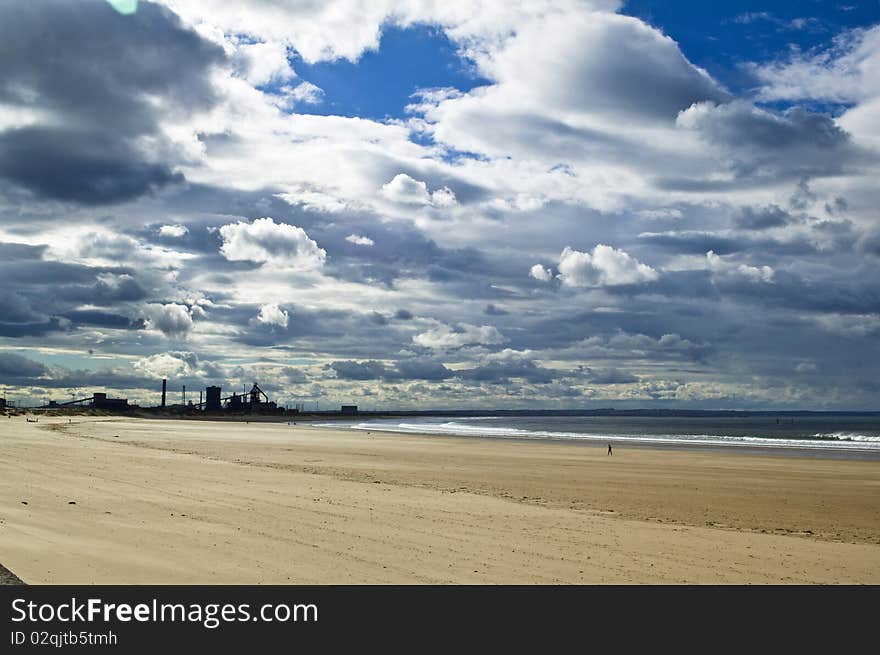 Lonely man walking on beach with industry in background and dramatic sky