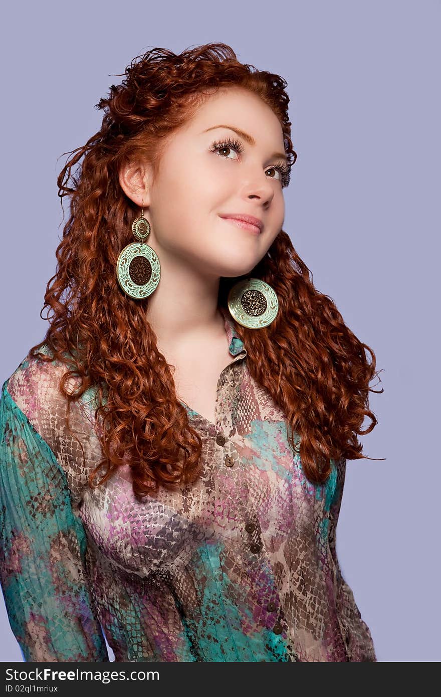 Relaxing and dreaming sensual red haired caucasian girl with positive and calm facial expression wearing extravagant round earrings and bright multicolored shirt standing isolated over blue background. Relaxing and dreaming sensual red haired caucasian girl with positive and calm facial expression wearing extravagant round earrings and bright multicolored shirt standing isolated over blue background