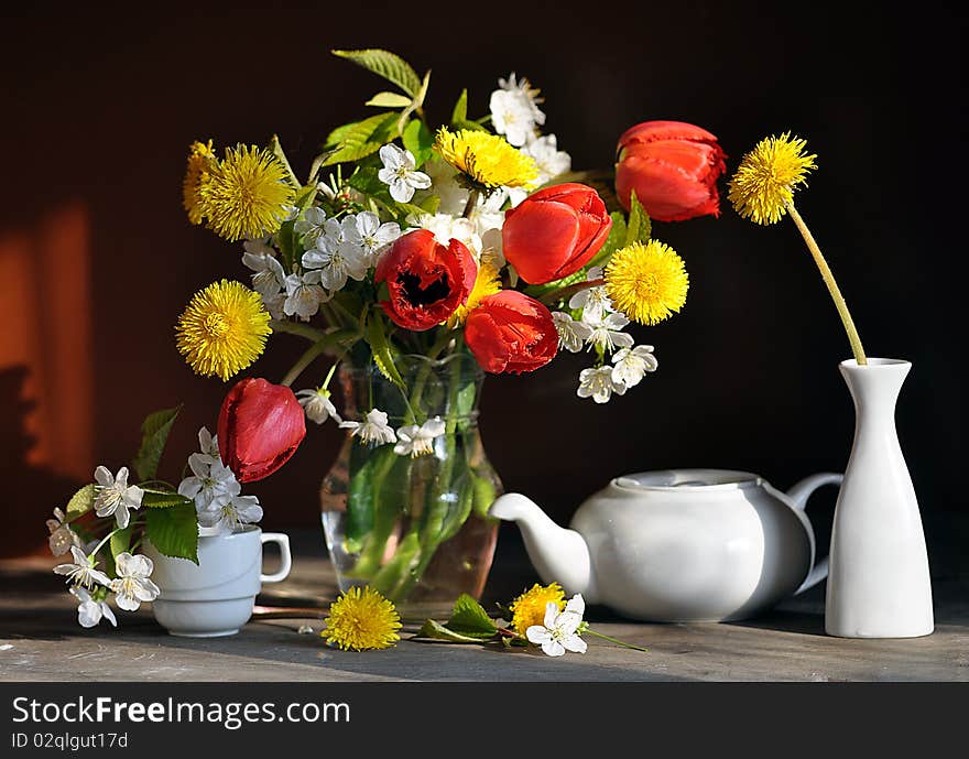 tulips, dandelions and branch of flowering cherry, stand in a glass vase, alongside tea-pot and cup. tulips, dandelions and branch of flowering cherry, stand in a glass vase, alongside tea-pot and cup