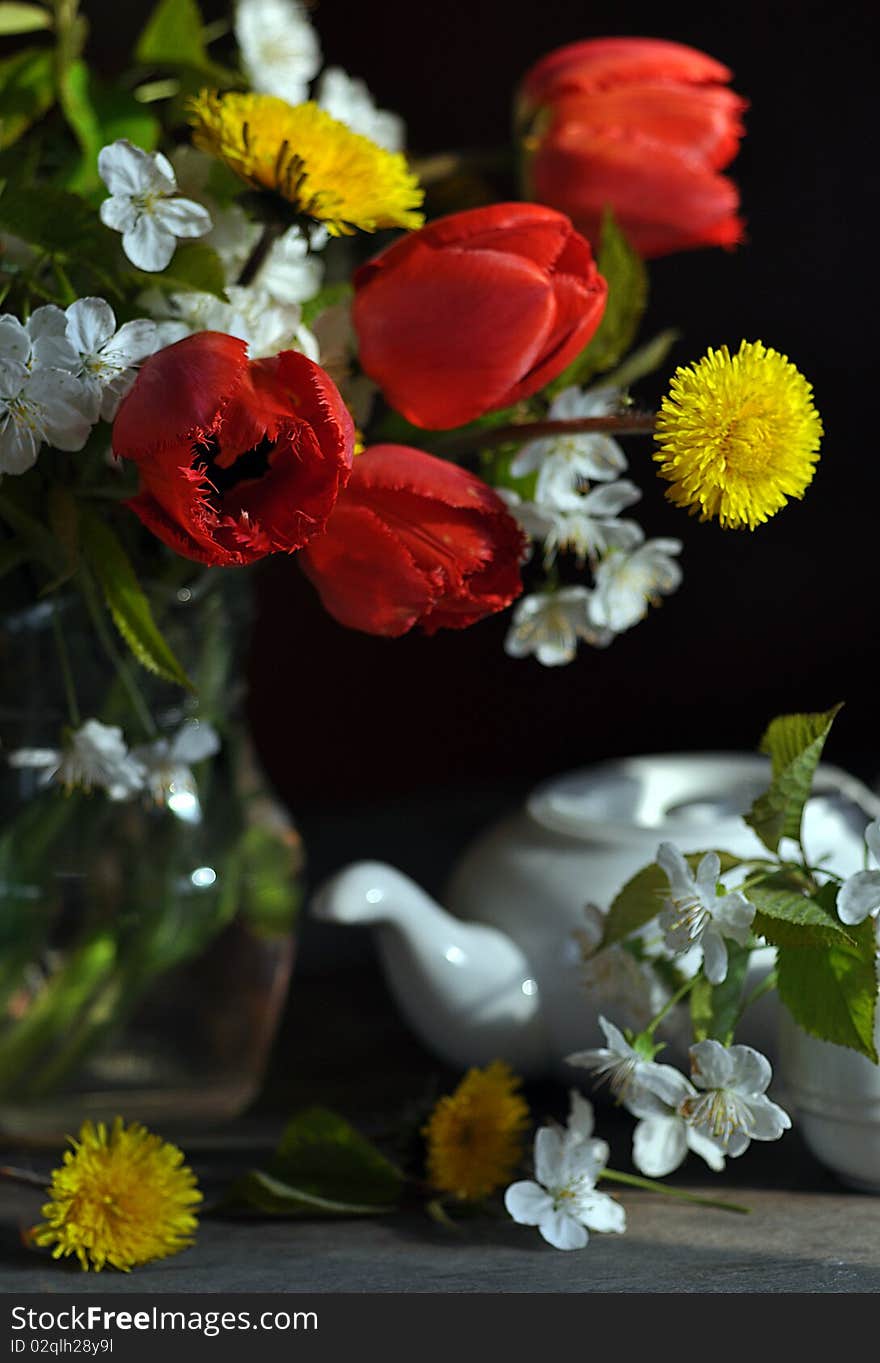 Tulips, dandelions and branch of flowering cherry, stand in a glass vase, alongside tea-pot and cup. Tulips, dandelions and branch of flowering cherry, stand in a glass vase, alongside tea-pot and cup