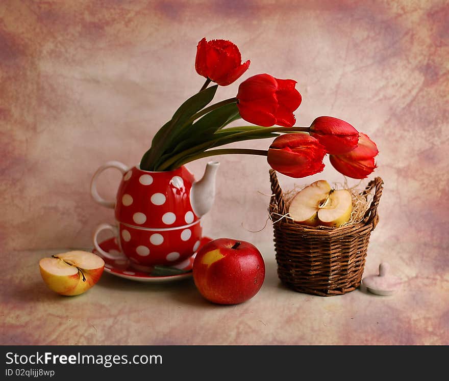 The bouquet of tulips stands in a tea-pot, alongside small basket and apples. The bouquet of tulips stands in a tea-pot, alongside small basket and apples