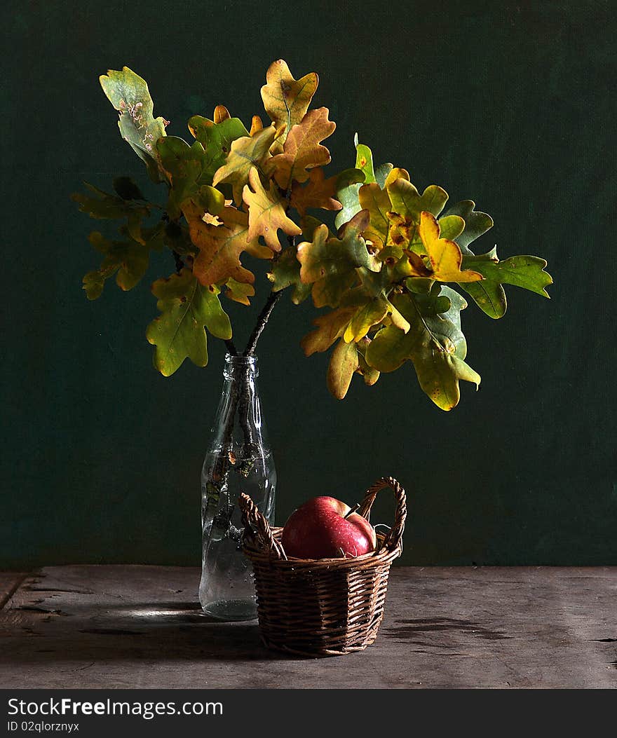 A branch with the leaves of oak stands in a glass bottle, alongside an apple lies in a small basket. A branch with the leaves of oak stands in a glass bottle, alongside an apple lies in a small basket