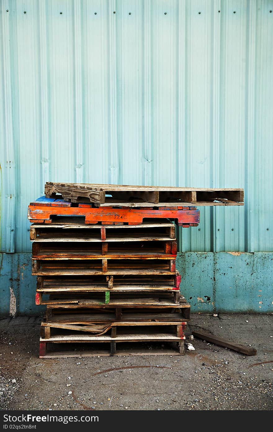 A stack of old wooden pallets painted in bright colors, next to the blue corrugated metal wall of an old factory building. A stack of old wooden pallets painted in bright colors, next to the blue corrugated metal wall of an old factory building.