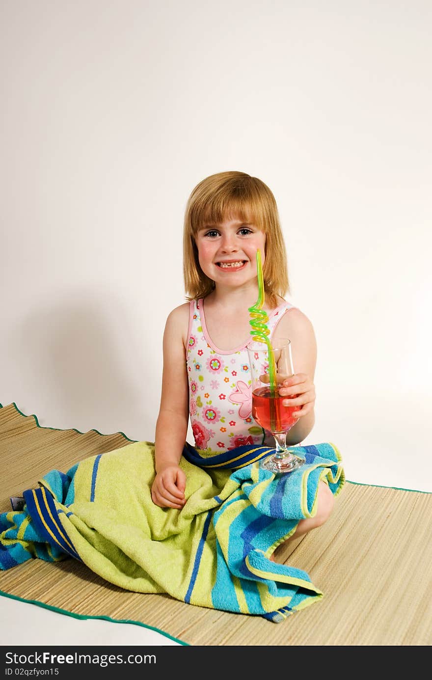 A vertical image of a lovely young girl in bathing costume holding a drink and sitting on a beach mat as she plays at being at the seaside,isolated against a plain background. A vertical image of a lovely young girl in bathing costume holding a drink and sitting on a beach mat as she plays at being at the seaside,isolated against a plain background