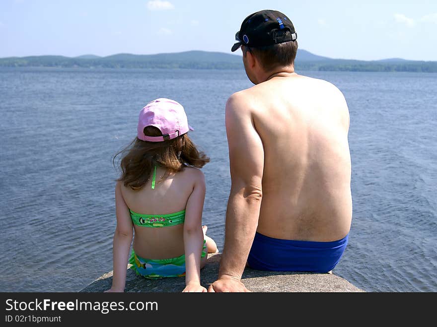 The man in swimming trunks and the girl in a bathing suit sit having turned a back ashore at water. The man in swimming trunks and the girl in a bathing suit sit having turned a back ashore at water