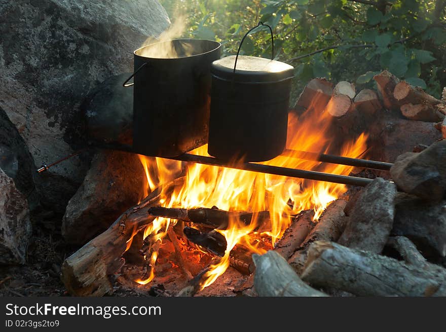 Two tourist kettles over campfire around rock with firewood