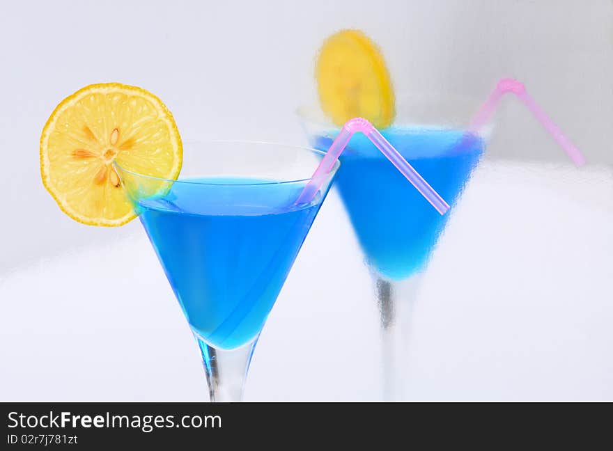 A blue cocktail with a lemon & a straw reflected in a silvery surface. A blue cocktail with a lemon & a straw reflected in a silvery surface.