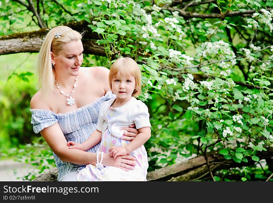 Woman and her baby daughter sit on a tree trunk in a beautiful blooming garden. Woman and her baby daughter sit on a tree trunk in a beautiful blooming garden