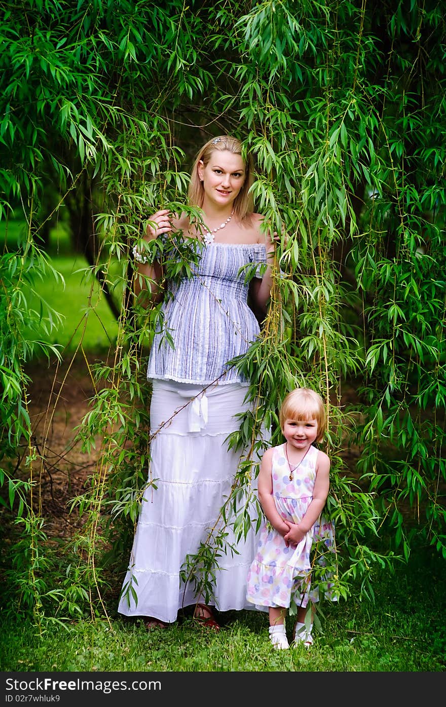 Beautiful pregnant woman and her adorable baby daughter stand in the willow garden. Beautiful pregnant woman and her adorable baby daughter stand in the willow garden
