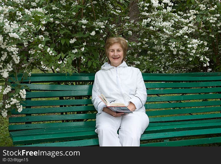 Elderly woman with a book in hands. Sits on a bench in a garden
Smiles and looks in a photographic camera