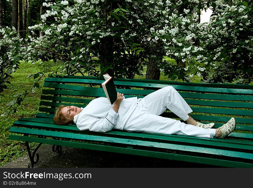 Ŭderly woman reads a book lying on a bench in a garden near a flowering bush