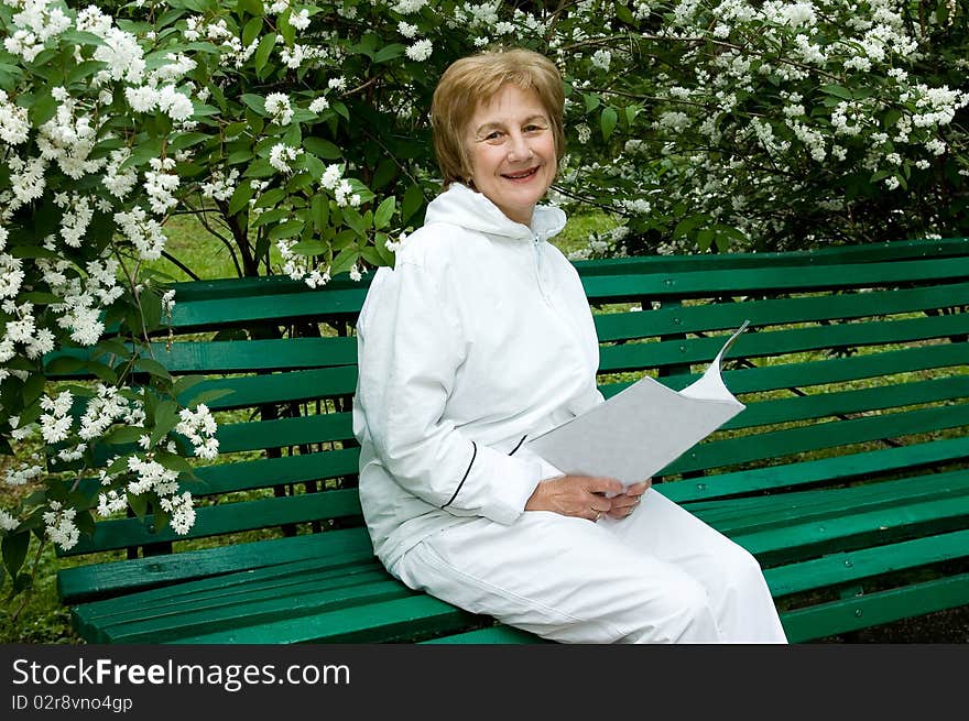 Elderly woman with a illustrated magazine in hands. Sits on a bench in a garden
Smiles and looks in a photographic camera