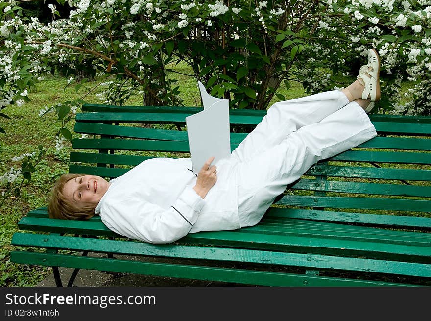 Elderly woman with a illustrated, magazine in hands.  
Rests on a bench in a garden.
Smiles and looks in a photographic camera