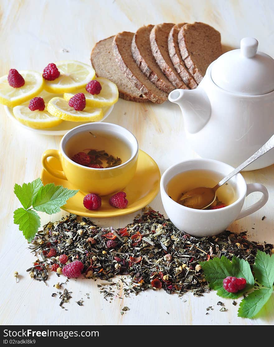 Two cups herbal tea is brewed in which, alongside tea-pot, lemon, bread and raspberry