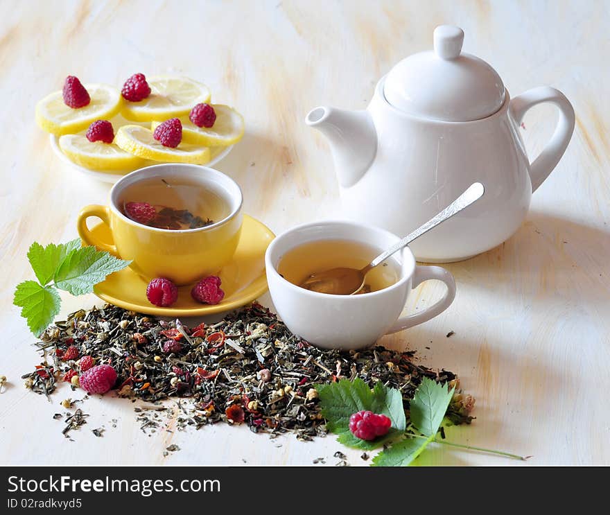 Two cups herbal tea is brewed in which, alongside tea-pot, lemon, and raspberry