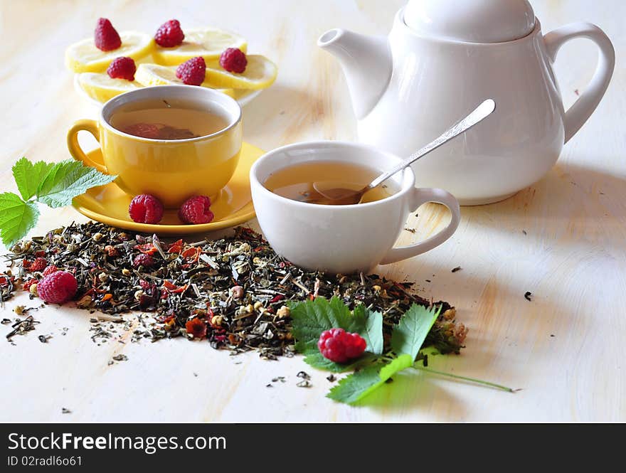 Two cups herbal tea is brewed in which, alongside tea-pot, lemon, and raspberry