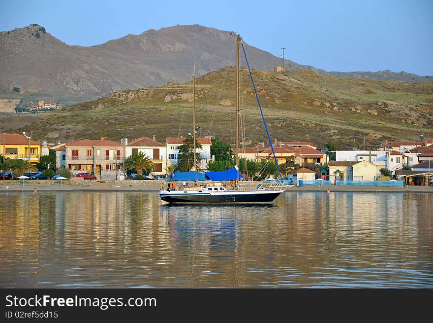 Dark blue sailboat reflected in calm sea water at sunset, in front of lovely small fishing village