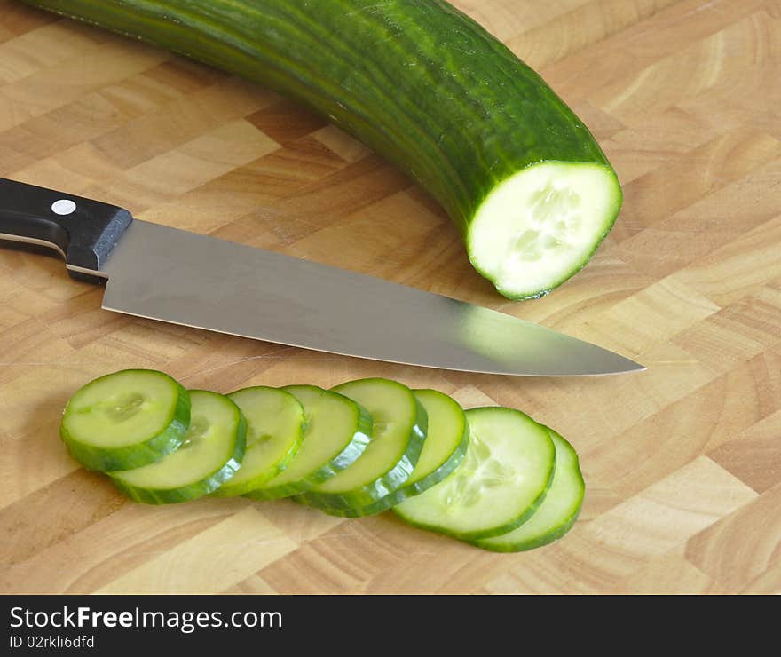 Kitchen knife rests on wooden chopping board with sliced cucumber.