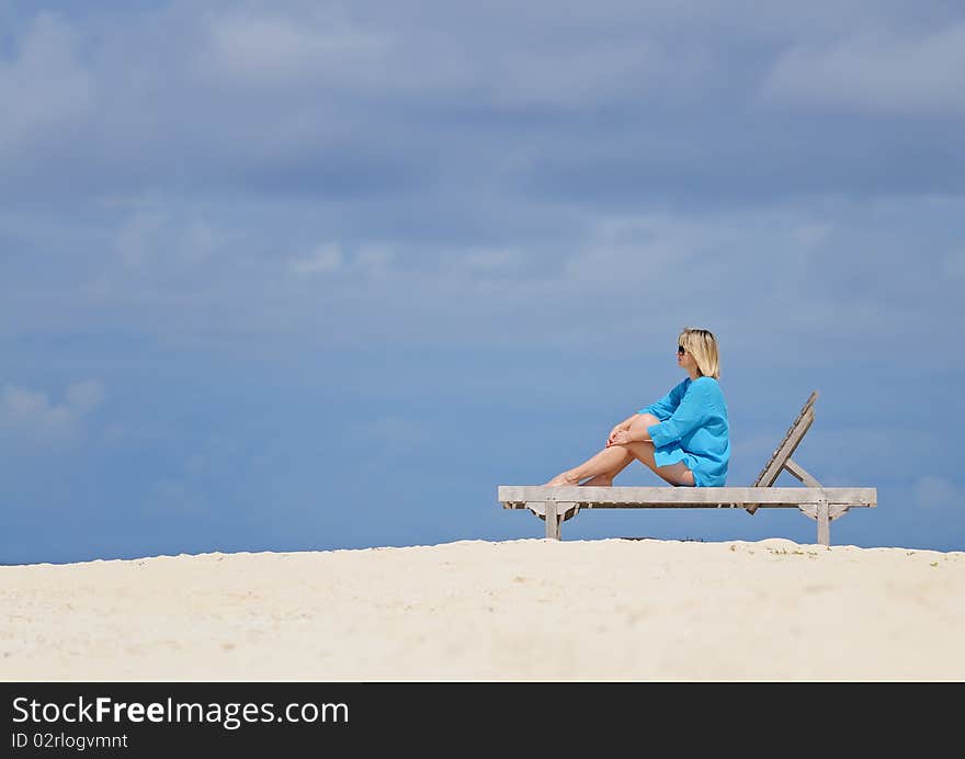 Woman in blue tunic sitting on wooden deck-chair on sandy beach against cloudy sky. Woman in blue tunic sitting on wooden deck-chair on sandy beach against cloudy sky.