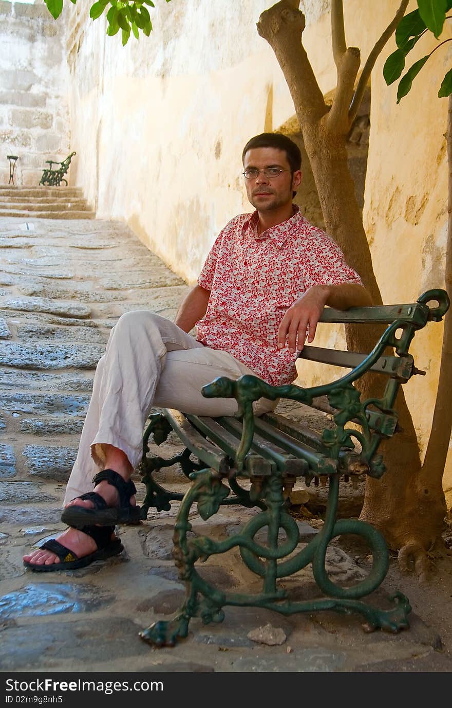 Young man sitting on old green bench under the tree, far from it there is another bench. Young man sitting on old green bench under the tree, far from it there is another bench