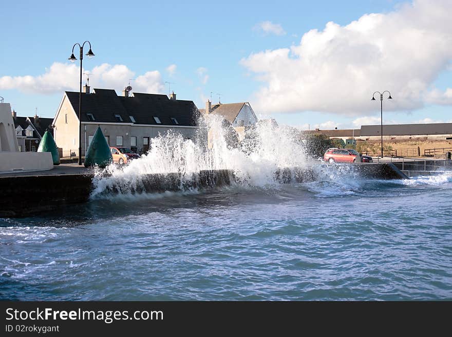 Waves splashing against a coastal wall during a storm with cars and homes in background. Waves splashing against a coastal wall during a storm with cars and homes in background