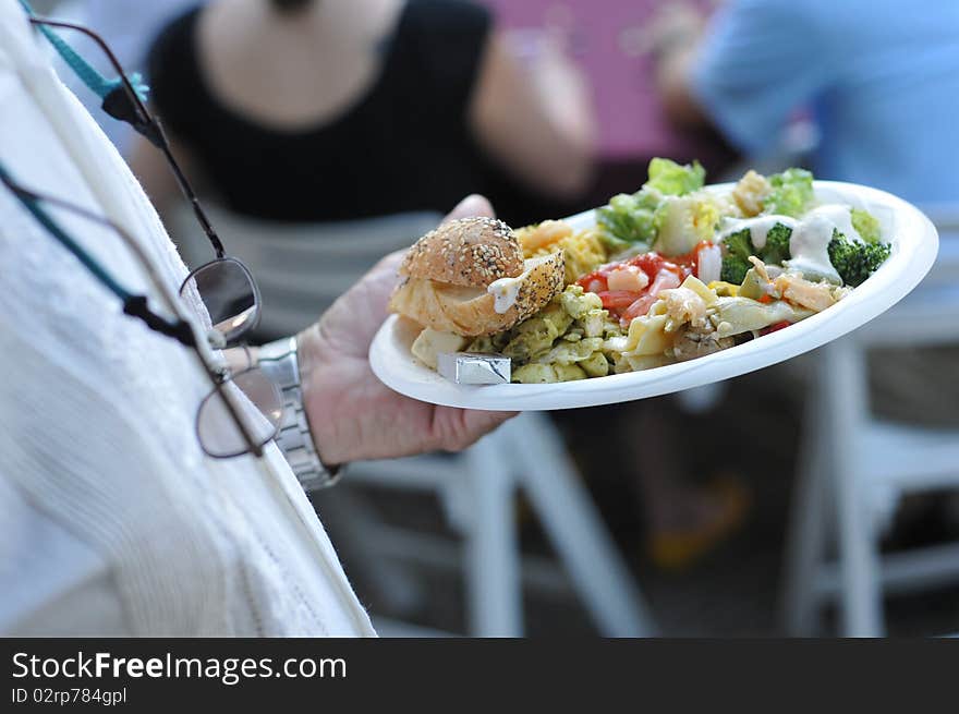 A man walks back to his table with his paper plate filled with food at a party. A man walks back to his table with his paper plate filled with food at a party.