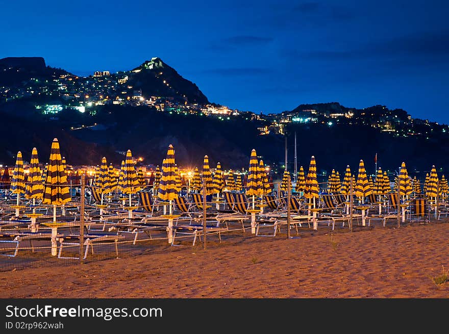 Beach at night with an illuminated village in the mountains and blue sky. The name of the village is Taormina, which is situated on Sicily, Italy.