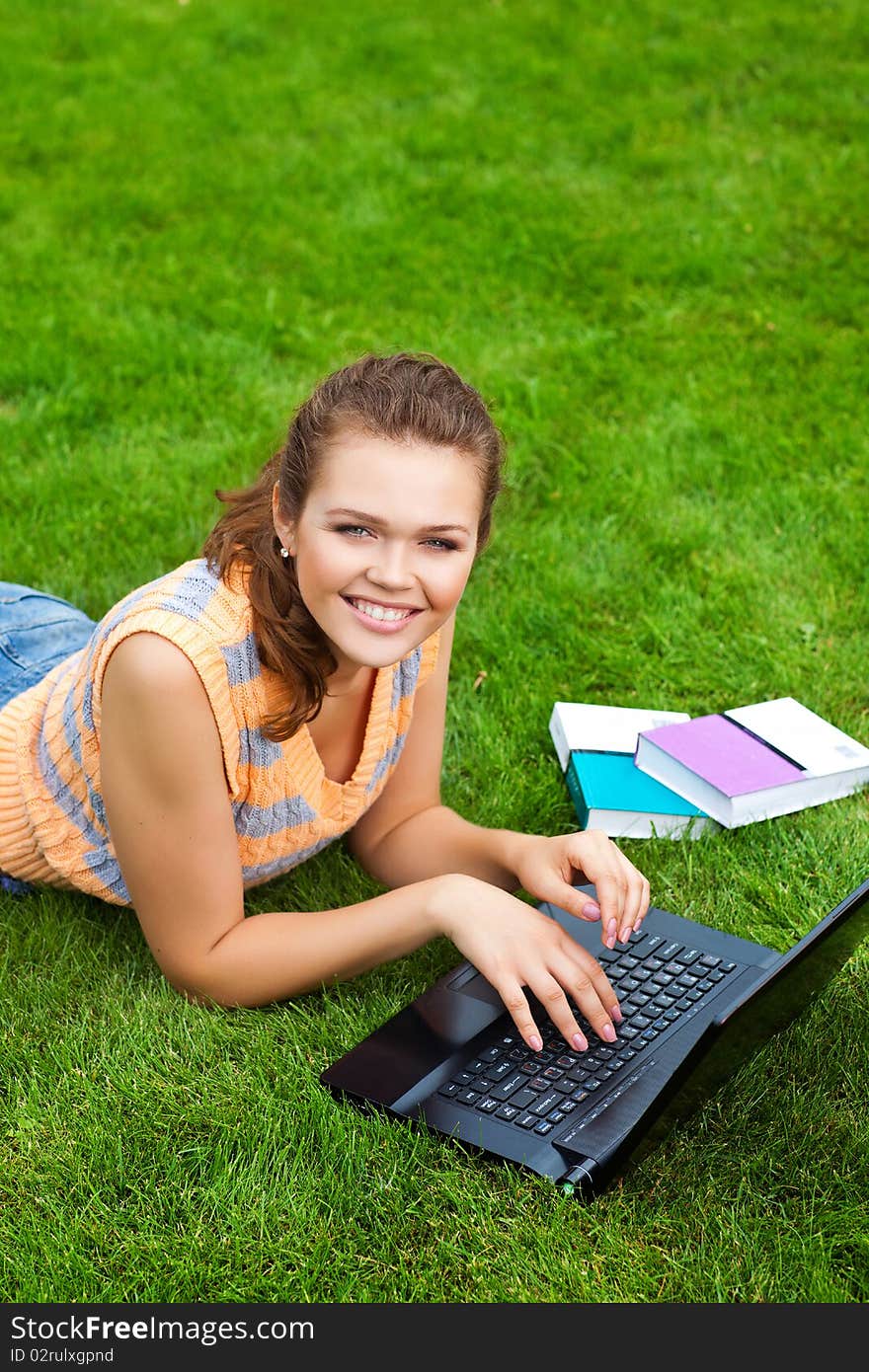 Pretty caucasian teenager lying down on green grass with laptop and books. Pretty caucasian teenager lying down on green grass with laptop and books