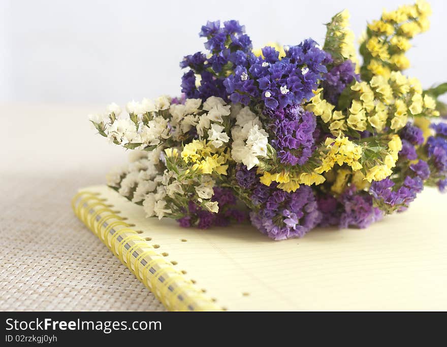 A bunch of violet,blue, yellow and white immortelles on a yellow copybook lying on a straw tabletop. A bunch of violet,blue, yellow and white immortelles on a yellow copybook lying on a straw tabletop