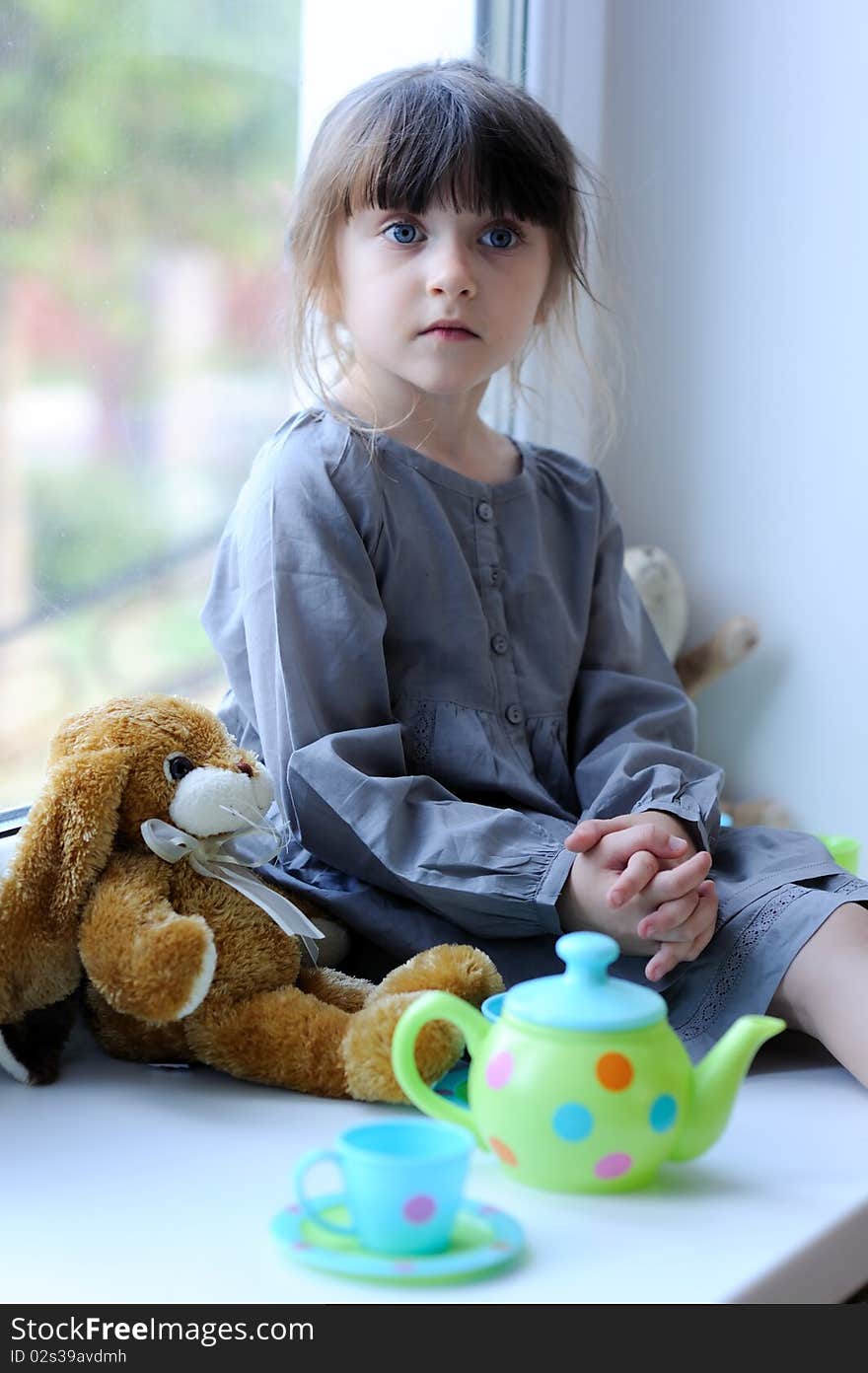 Nice toddler girl with toy tea set and bunny sitting near the winow in cute gray dress