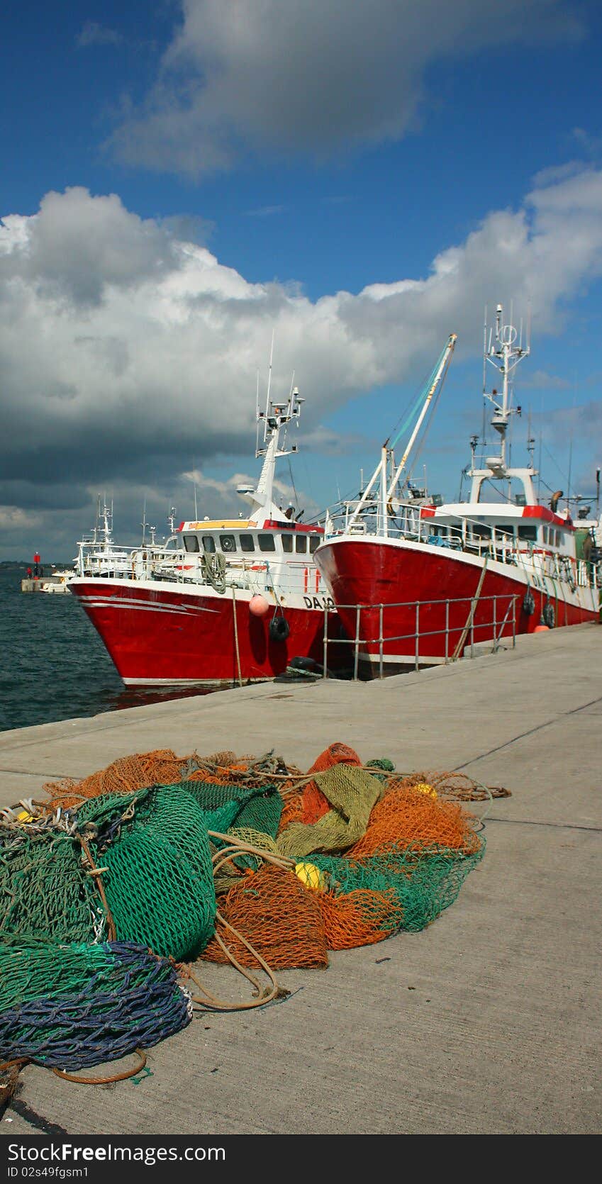 Green and blue and orange fishing nets stacked on a quayside in Ireland. The nets are pictured in front red and white trawlers. The sky is a rich blue with cumulus clouds. Green and blue and orange fishing nets stacked on a quayside in Ireland. The nets are pictured in front red and white trawlers. The sky is a rich blue with cumulus clouds.