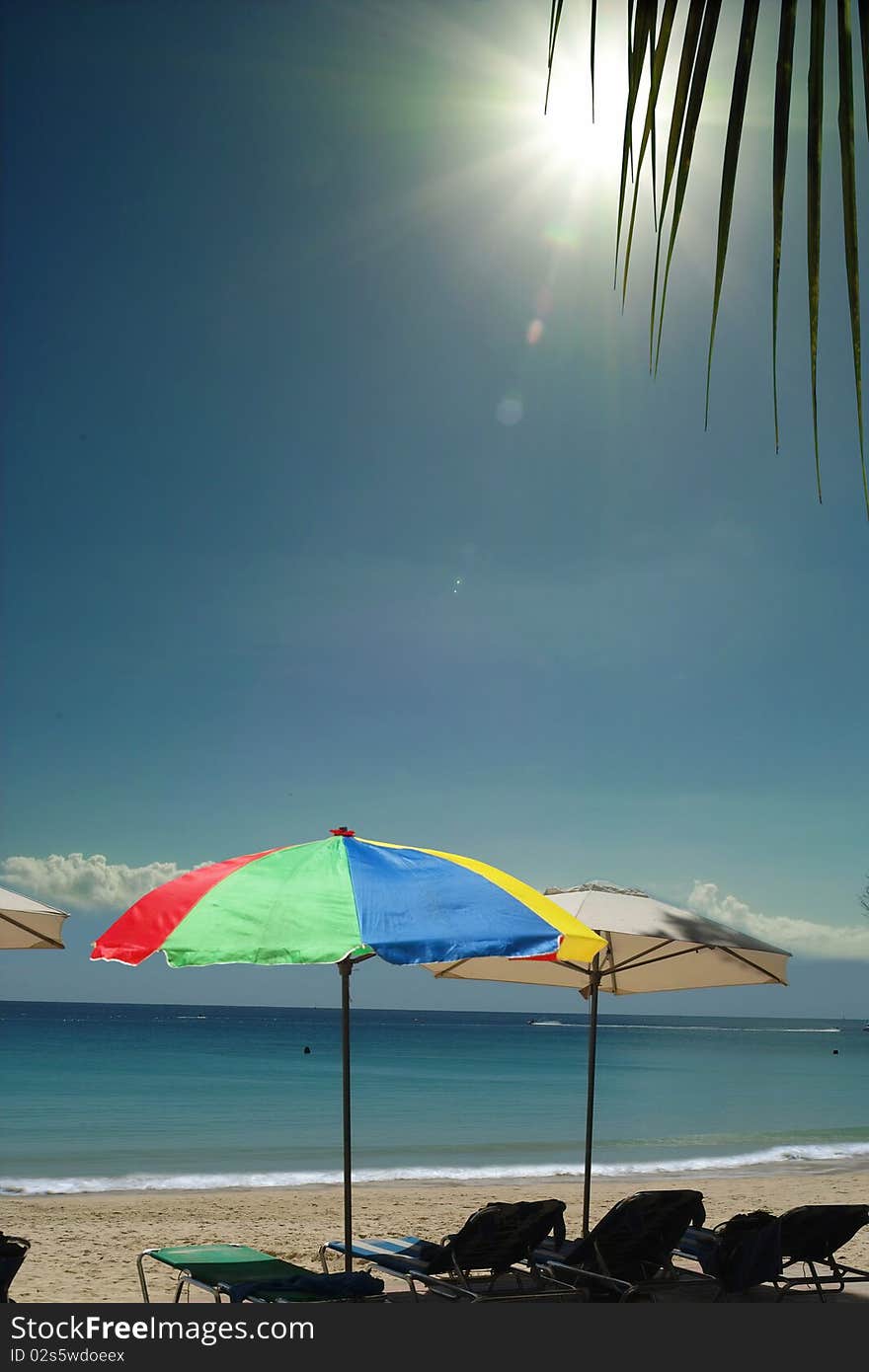 The colorful umbrellas, chairs are waiting on the tropical sea beach. Sun is shining above on the blue sky trough the leaf of palm-tree. The sky area is free for your text. The colorful umbrellas, chairs are waiting on the tropical sea beach. Sun is shining above on the blue sky trough the leaf of palm-tree. The sky area is free for your text.