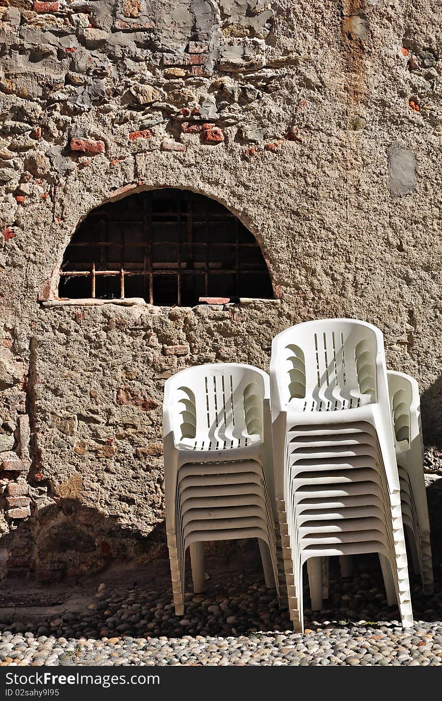 Some stacks of modern white plastic chairs with an old wall like background in a street of Garessio, in Piedmont, Italy.