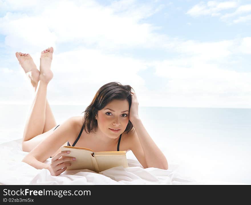 A young and sexy brunette is lying on a white silk blanket and reading a book. The image is taken outdoors, on a background with sky and water. A young and sexy brunette is lying on a white silk blanket and reading a book. The image is taken outdoors, on a background with sky and water.