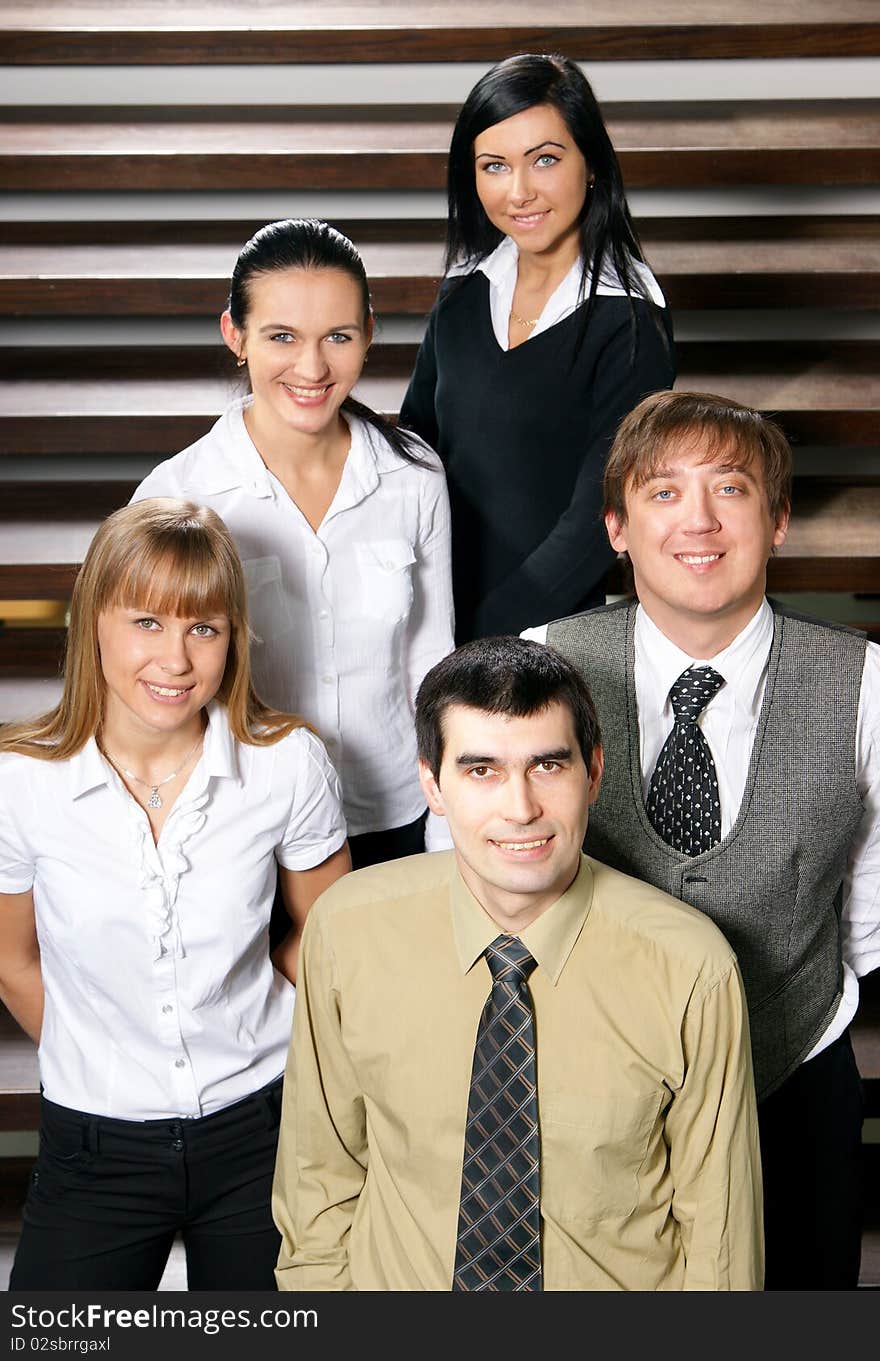 Five young business persons are standing as a team with a man in front. The image is taken on a background with stairs. Five young business persons are standing as a team with a man in front. The image is taken on a background with stairs.