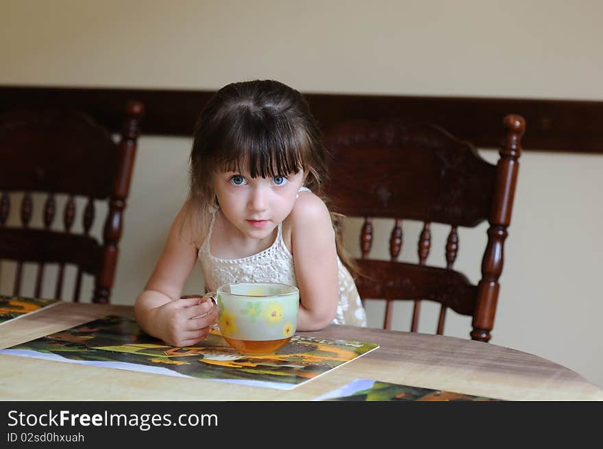 Nice toddler girl with big cup of tea sitting near the table and looking into the camera
