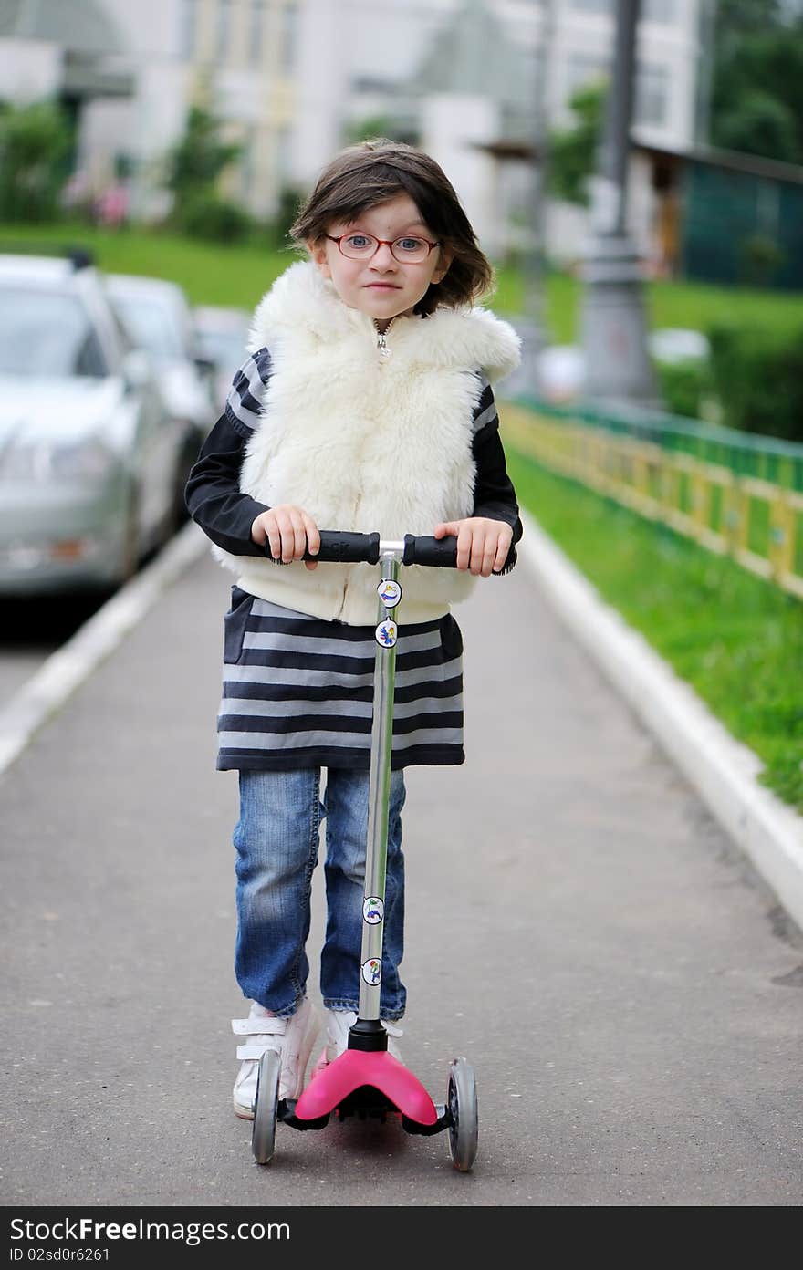 Nice little toddler girl in stripe dress, jeans and white fur jacket riding on her pink scooter. Nice little toddler girl in stripe dress, jeans and white fur jacket riding on her pink scooter