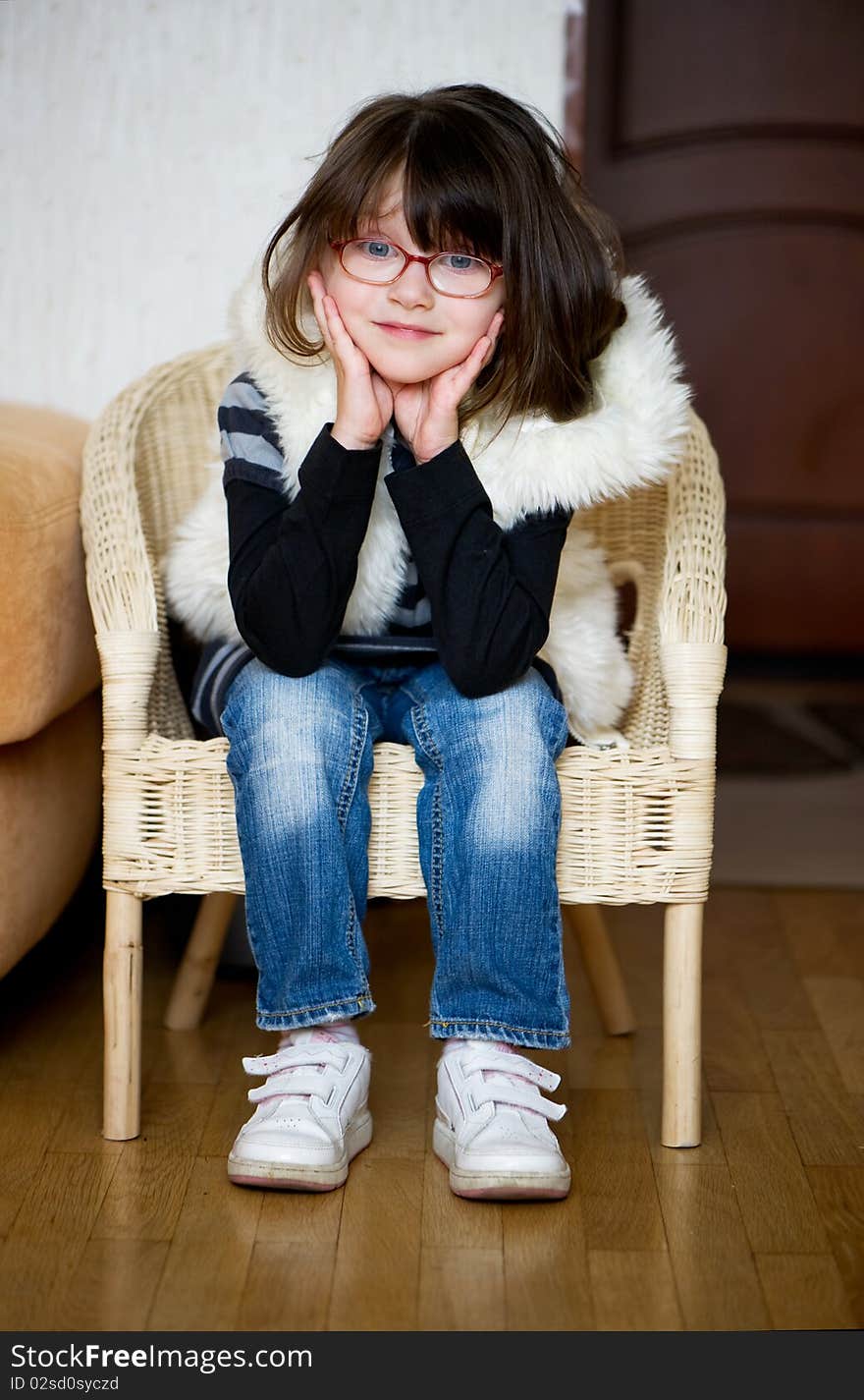 Nice toddler girl in glasses and white fur jacket with dark hair sitting in chair and looking to the camera. Nice toddler girl in glasses and white fur jacket with dark hair sitting in chair and looking to the camera