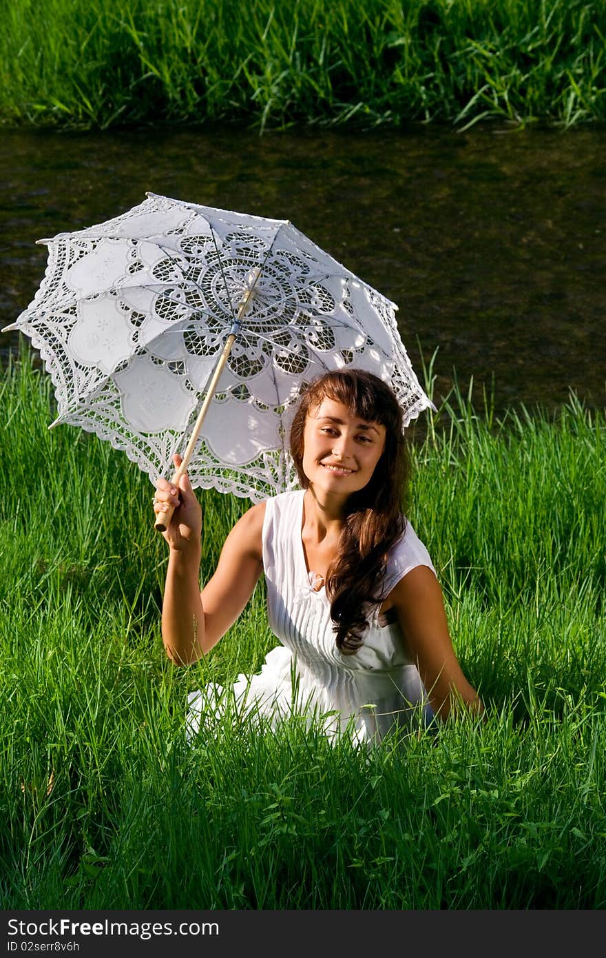 Young  attractive girl in white with sun umbrella sitting in the fresh green grass near the river. Young  attractive girl in white with sun umbrella sitting in the fresh green grass near the river