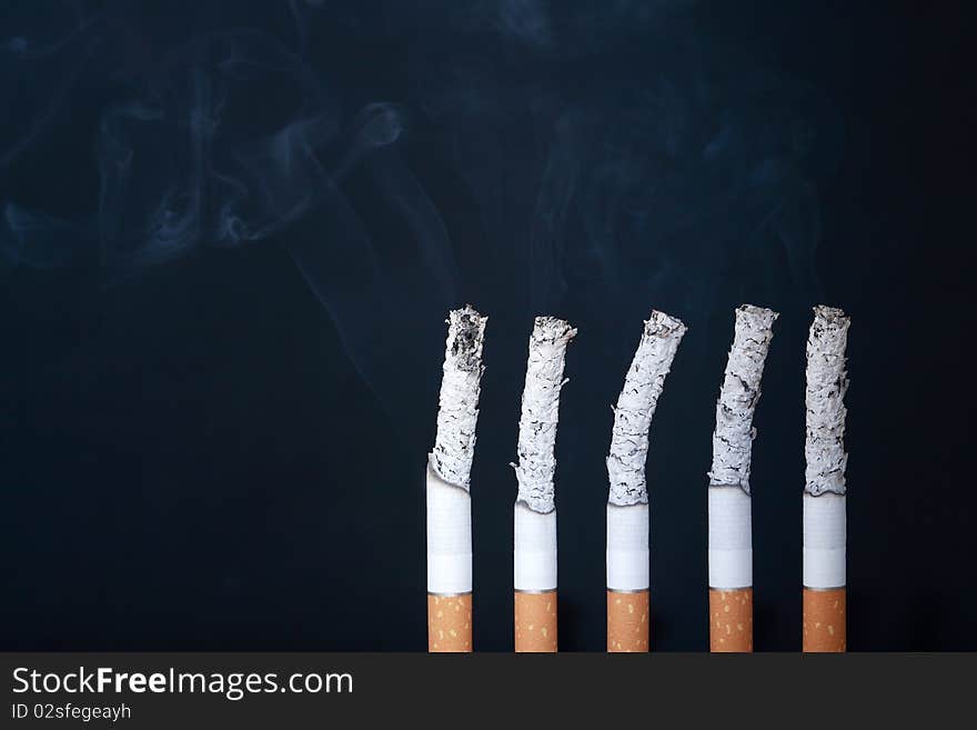 Five cigarettes with smoke and ashes on dark background