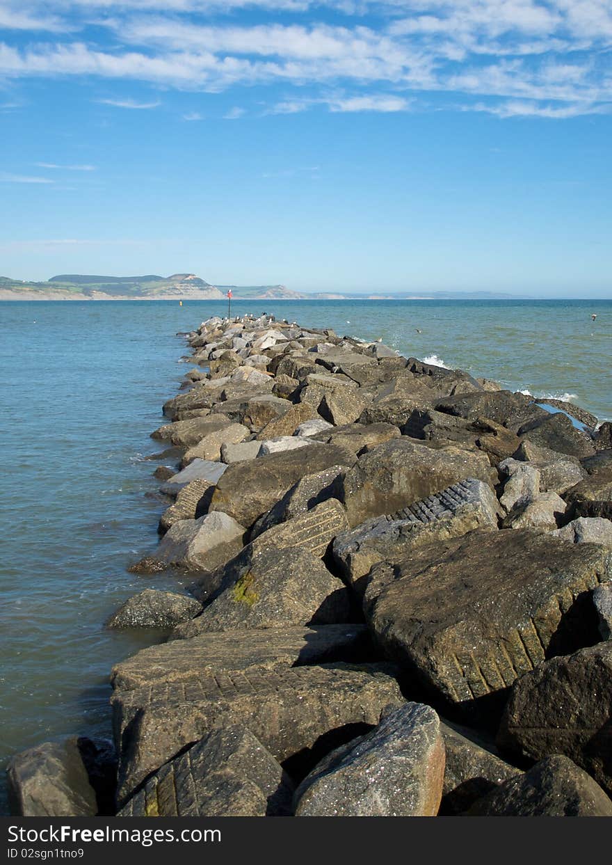 A stone water brake stretching out across the bay. Cliffs in the background with whispery clouds in the sky. Was taken in Lyme Regis, UK