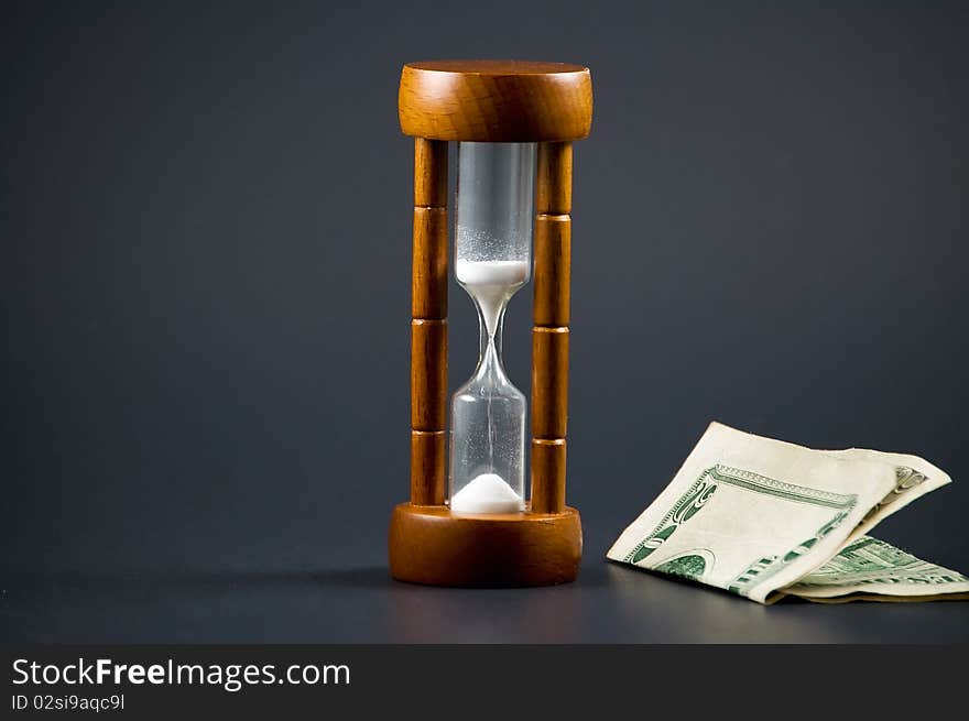 Folded twenty dollar bill is seen next to a wooden hourglass against black background. This image is  a metaphor for the saying  Time is Money
