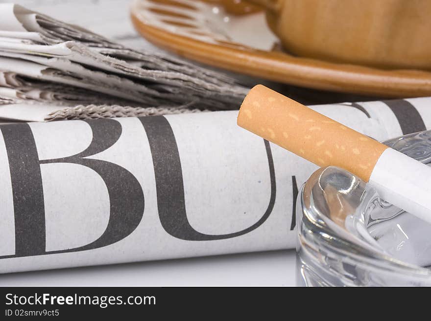 A filtered cigarette resting on a glass ashtray next to a brown cup and plate and a newspaper.