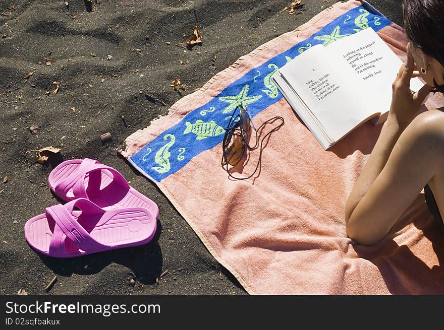 Beach scene with a partial view of a woman lying on an orange beach towel reading a book in the right side and a pair of pink sandals on the left; horizontal view