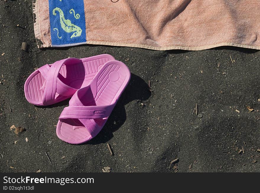 Beach scene with a partial view of an orange beach towel and a pair of pink sandals on the volcanic black sand; horizontal view. Beach scene with a partial view of an orange beach towel and a pair of pink sandals on the volcanic black sand; horizontal view